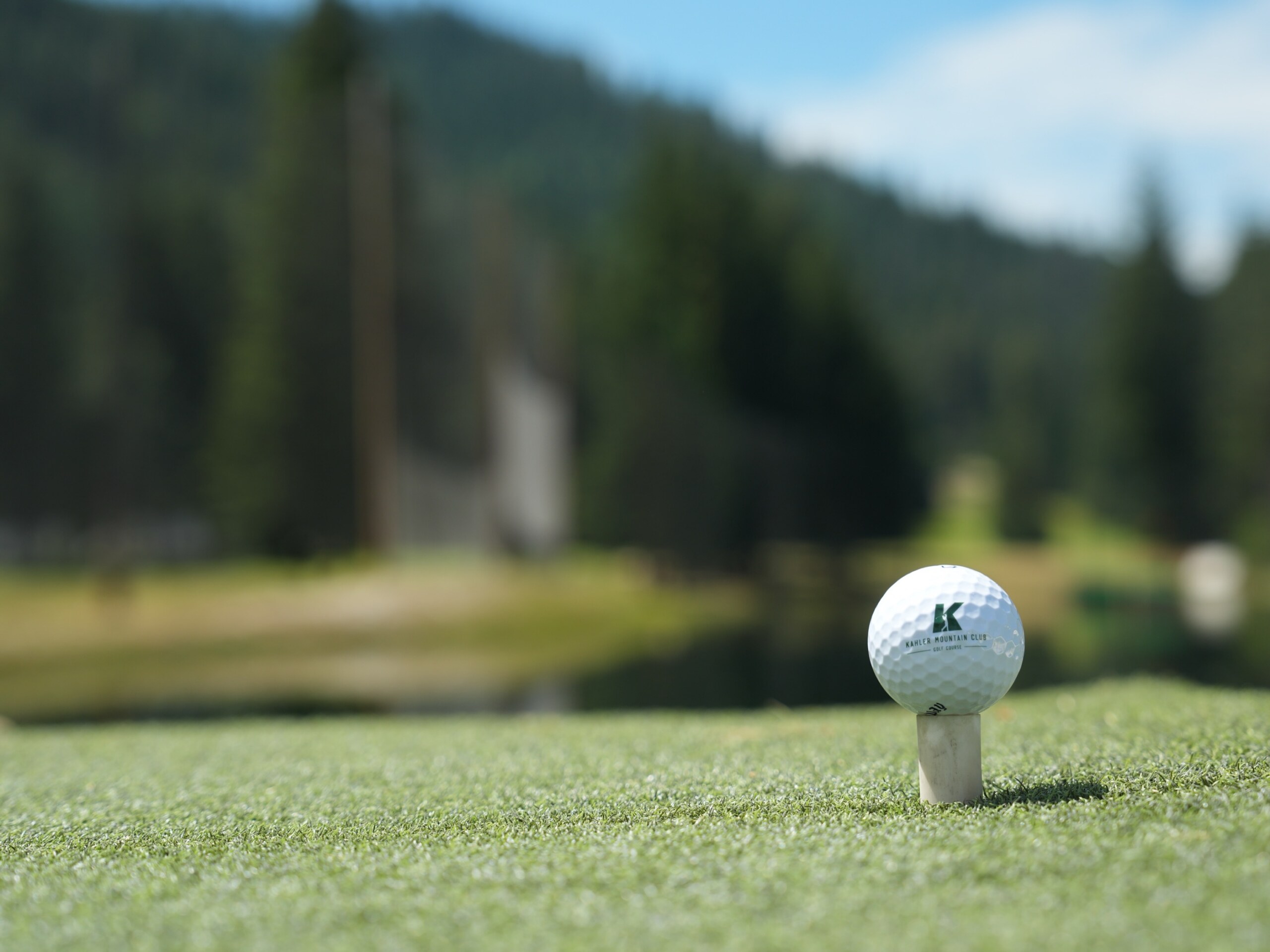 A golf ball carrying the Kahler Mountain Club logo, teed up with the water and mountains behind.